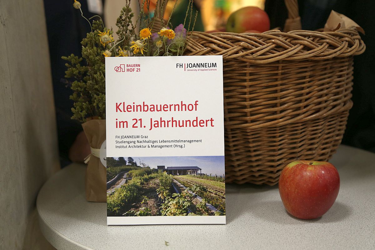 Folder zur Veranstaltung "Kleinbauernhof im 21. Jahrhundert", im Hintergrund ein Korb mit Äpfeln.