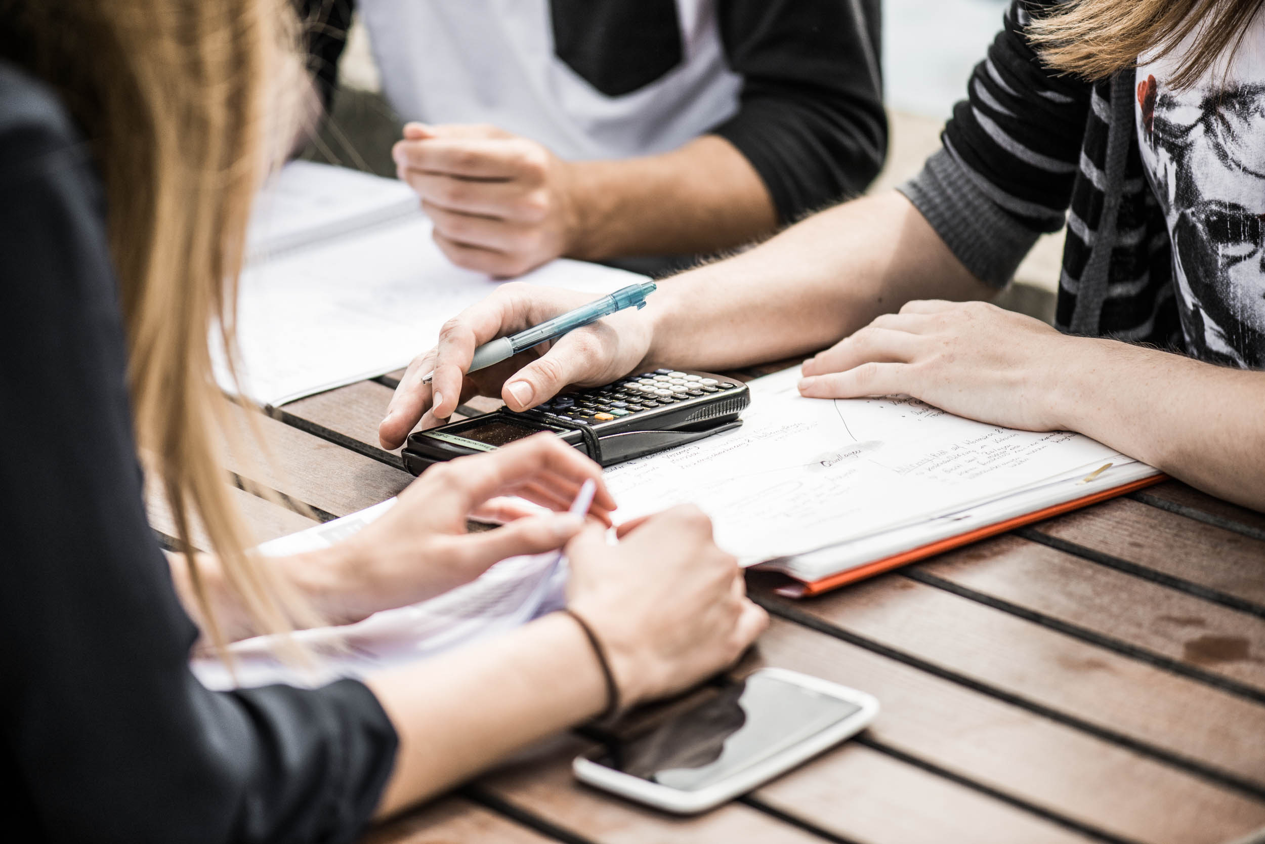 Studierende sitzen mit Block, Stift, Taschenrechner und Smartphone an einem Holztisch.