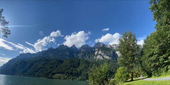 Berge mit grünen Hängen und Felsen unter blauem Himmel mit Wolken, daneben ein See.