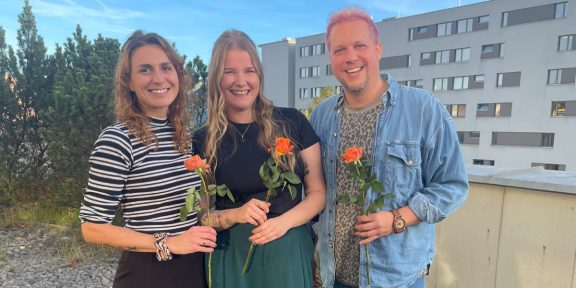 Drei Personen, zwei Frauen und ein Mann mit pinken Haaren, halten Rosen in der Hand.